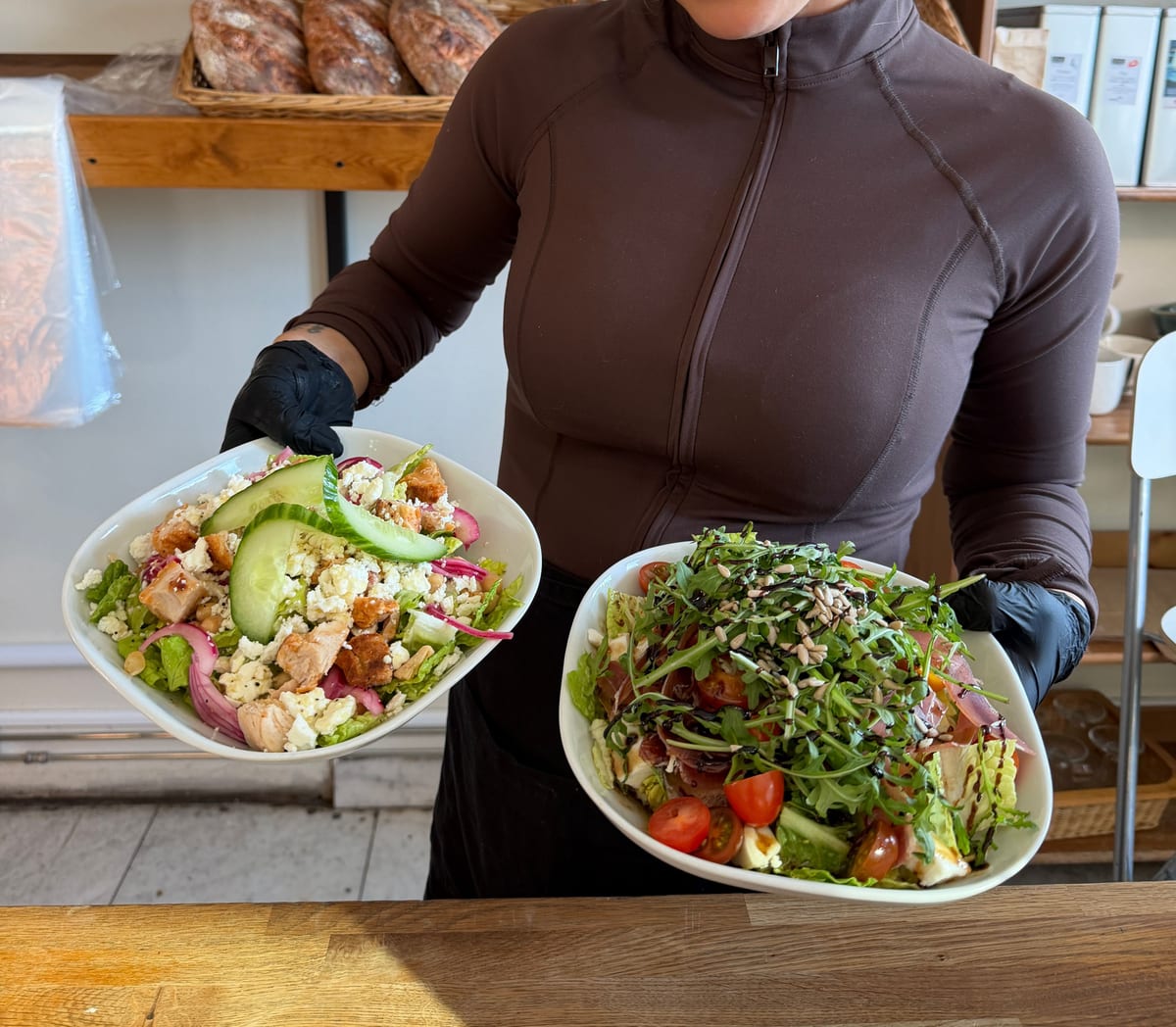 Två sallader hålls fram över disken, en med kyckling och fetaost och en med ruccola, körsbärstomater och frön